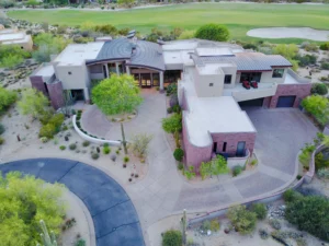 An aerial view of a modern, expansive home set within a desert landscape, featuring clean lines, multiple flat roofs, and a circular driveway. The property is surrounded by sparse desert vegetation, with a golf course visible in the background, highlighting the home’s secluded and luxurious setting. The architecture incorporates various textures and materials, blending with the natural surroundings.