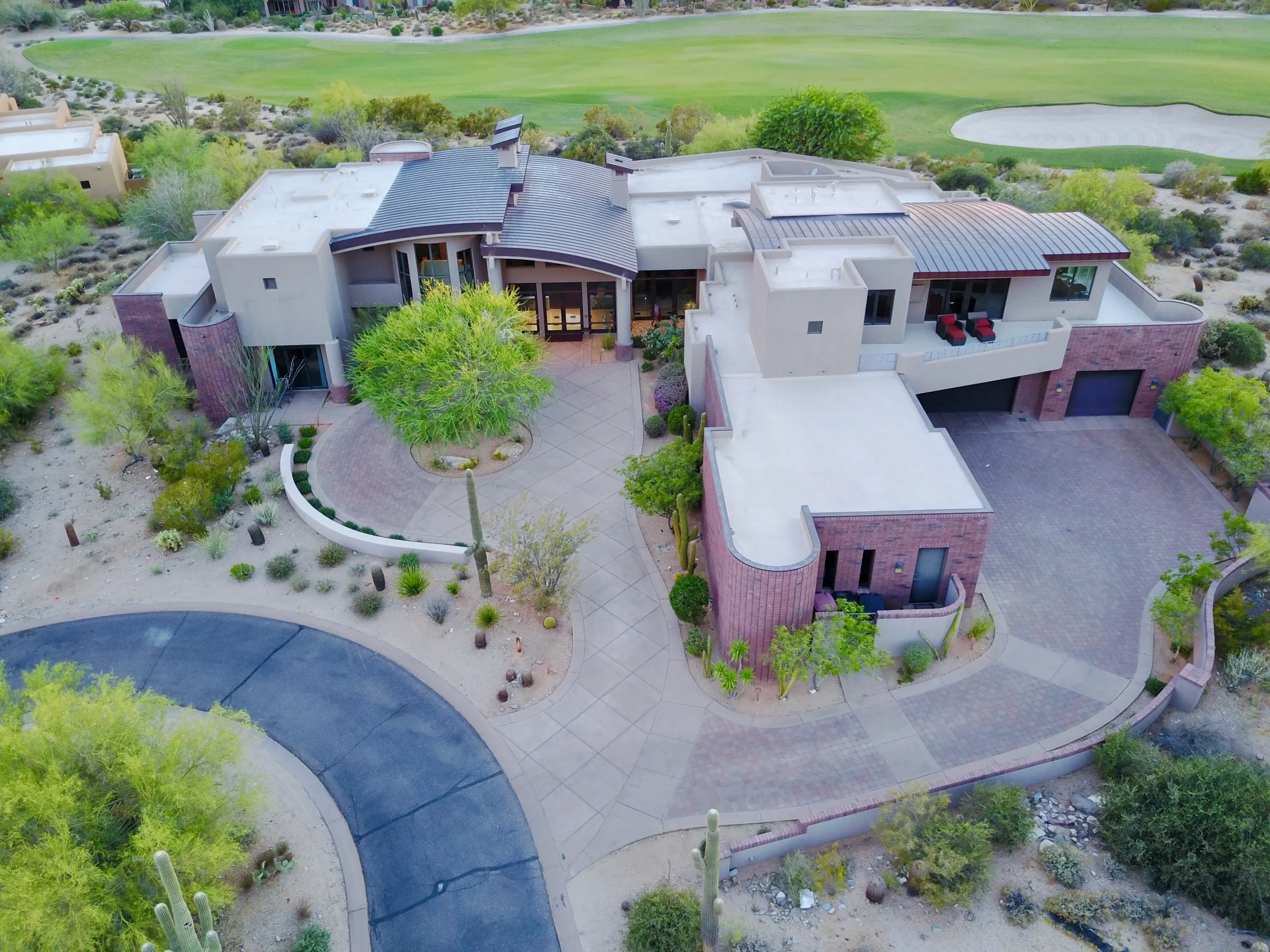 An aerial view of a modern, expansive home set within a desert landscape, featuring clean lines, multiple flat roofs, and a circular driveway. The property is surrounded by sparse desert vegetation, with a golf course visible in the background, highlighting the home’s secluded and luxurious setting. The architecture incorporates various textures and materials, blending with the natural surroundings.