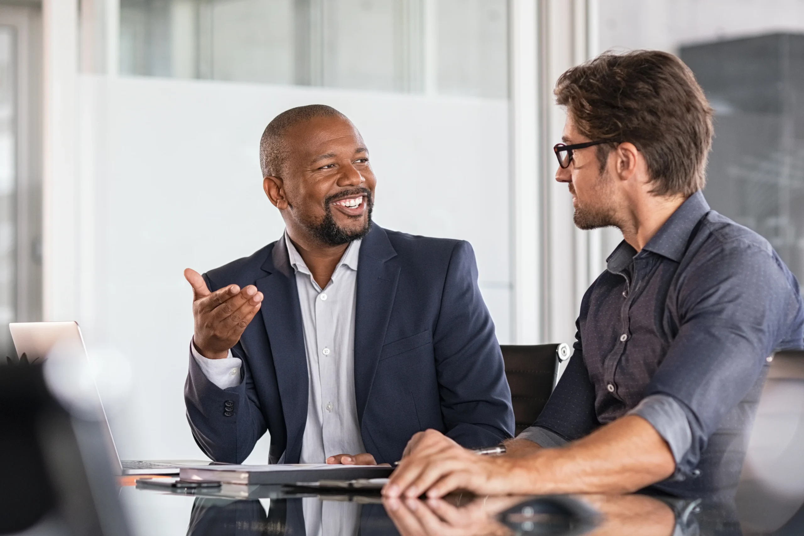 Two businessmen sit at a conference table engaged in a friendly discussion, with one gesturing expressively and smiling while the other listens attentively. Both appear professional and relaxed, fostering a collaborative atmosphere in a modern office setting. A laptop and documents are visible on the table, indicating a work-related conversation.