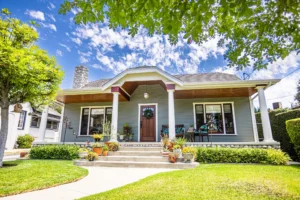A charming bungalow with a covered front porch, columns, and a neatly landscaped yard is shown under a bright blue sky. The home's exterior features a welcoming wreath on the wooden front door, potted plants lining the steps, and well-maintained greenery. The surrounding trees and open sky add to the inviting, serene atmosphere of this suburban home.