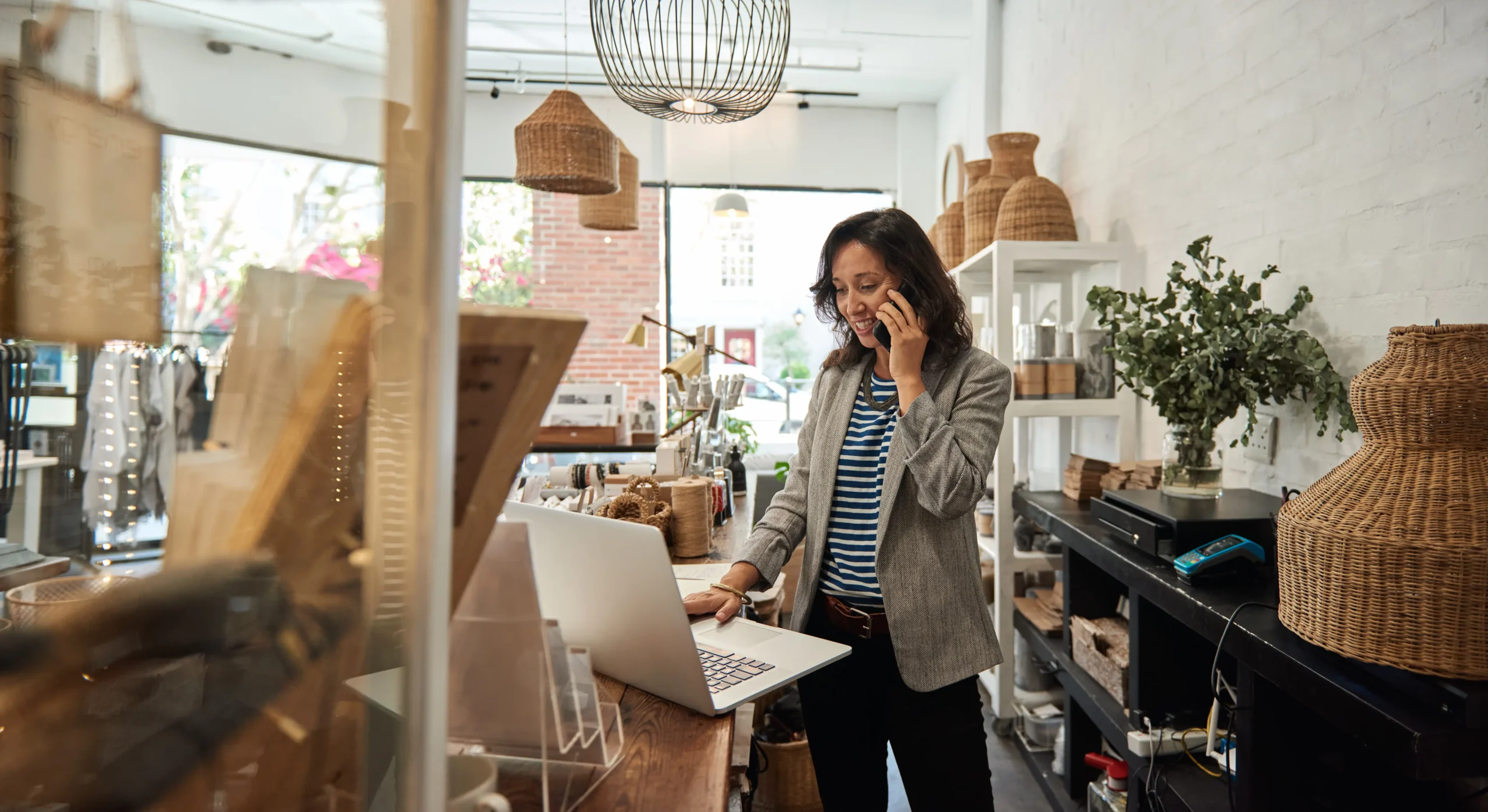 A woman in a small retail shop speaks on the phone while working on a laptop. She is surrounded by wicker baskets, shelves with decorative items, and a leafy plant, giving the space a cozy and artisan atmosphere. The bright natural light from the window highlights the shop's rustic decor.