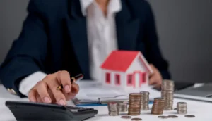 Businessperson conducting real estate financial planning with calculator, stacked coins, miniature house model, and business documents on desk.