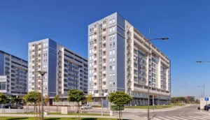Modern high-rise residential apartment complex in an urban setting, featuring contemporary architectural design under a clear blue sky, with city streetscape, parking signage, and landscaping.