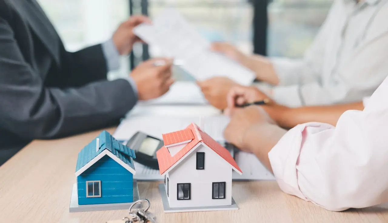 Business professionals discussing mortgage or property financing in a meeting, with miniature house models, a calculator, and financial documents on the table.